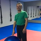 José-López una persona en un gimnasio o estudio de artes marciales con equipo de entrenamiento de suspensión visible al fondo. La persona sostiene una pelota de ejercicio o fitball azul grande,
